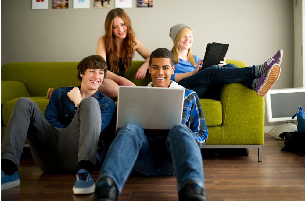 four smiling teens sitting on and around a couch looking at laptops