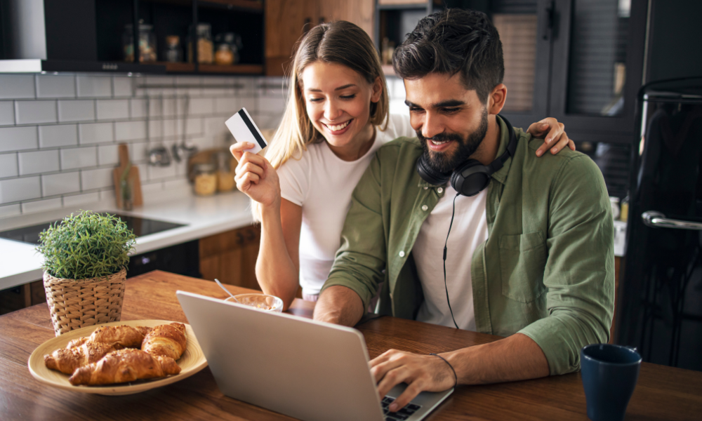 young couple sitting at kitchen counter with a laptop. He's typing, she's holding the credit card.