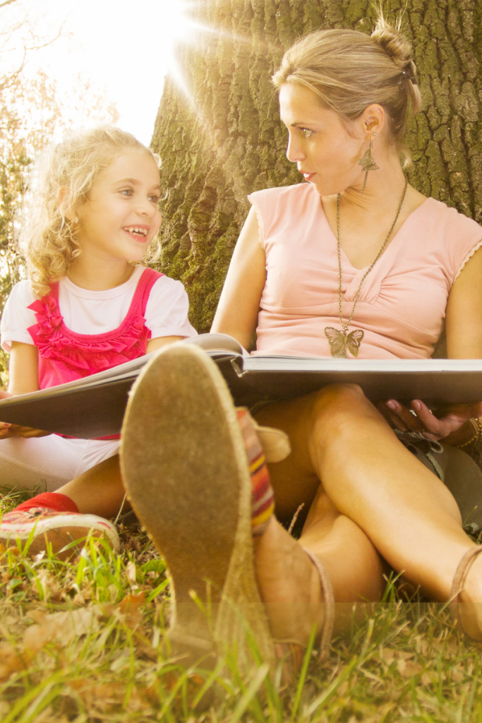 Mom and daughter sitting under a tree reading a book together in the sunlight